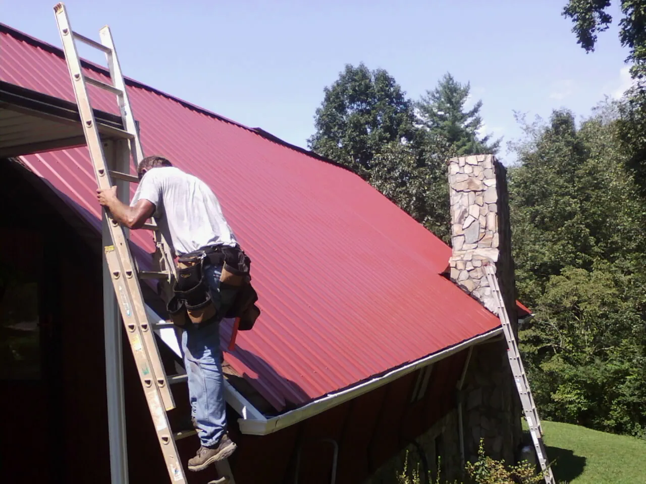 Roofer on a ladder beside a red metal roof and stone chimney — Soffit & Fascia Repair by Roofing Brothers in Whittaker